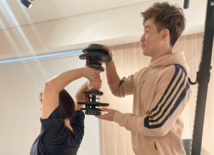 30代以降の女性がパーソナルトレーナーのサポートを受けながら、二の腕の引き締めを目的とした、二の腕のトレーニングを行っている様子。